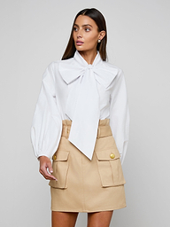 A woman stands against a plain background, wearing the Deon Tie-Neck Blouse—an organic cotton poplin top with blouson sleeves and an oversized neck tie—paired with a beige skirt featuring front pockets and gold buttons. Her long, straight hair frames her face as she gazes slightly to the side.