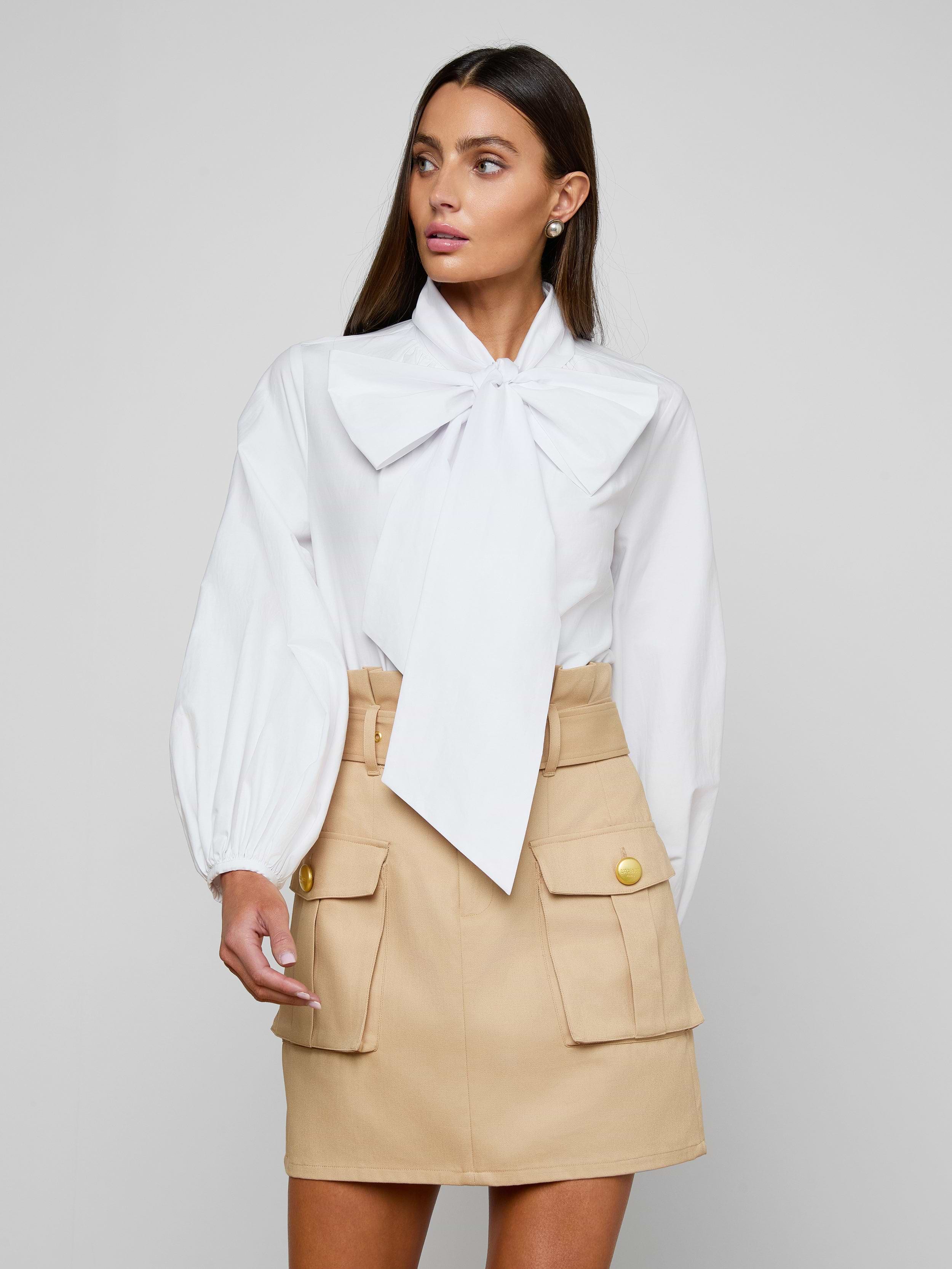 A woman stands against a plain background, wearing the Deon Tie-Neck Blouse—an organic cotton poplin top with blouson sleeves and an oversized neck tie—paired with a beige skirt featuring front pockets and gold buttons. Her long, straight hair frames her face as she gazes slightly to the side.