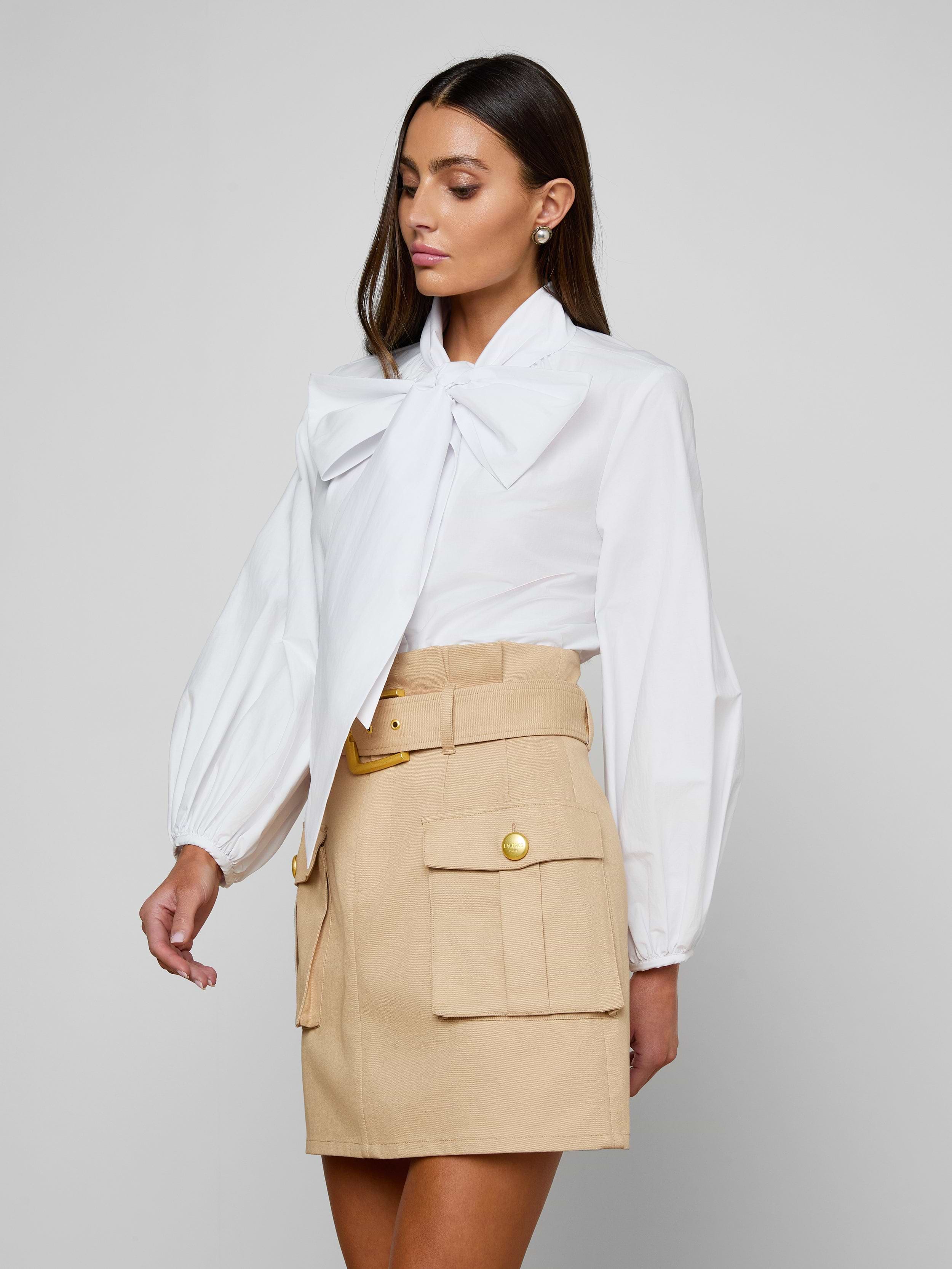 A woman is wearing the Deon Tie-Neck Blouse—a white organic cotton poplin top with blouson sleeves and an oversized neck tie—paired with a beige high-waisted skirt featuring front pockets and gold buttons, standing against an off-white background.
