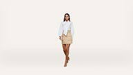 A woman walks forward wearing a white blouse, beige skirt, and nude heels against a plain white background.