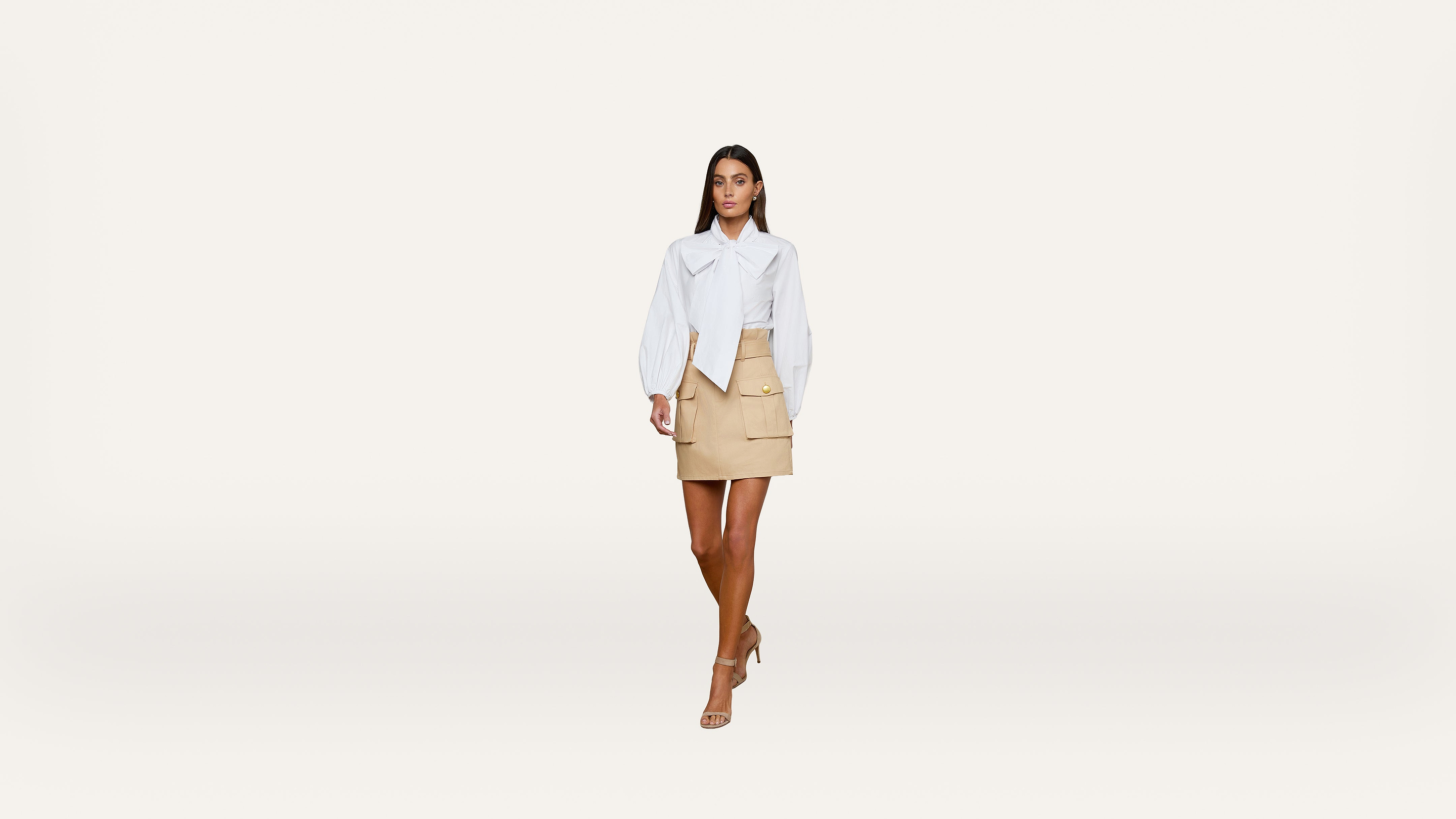 A woman walks forward wearing a white blouse, beige skirt, and nude heels against a plain white background.