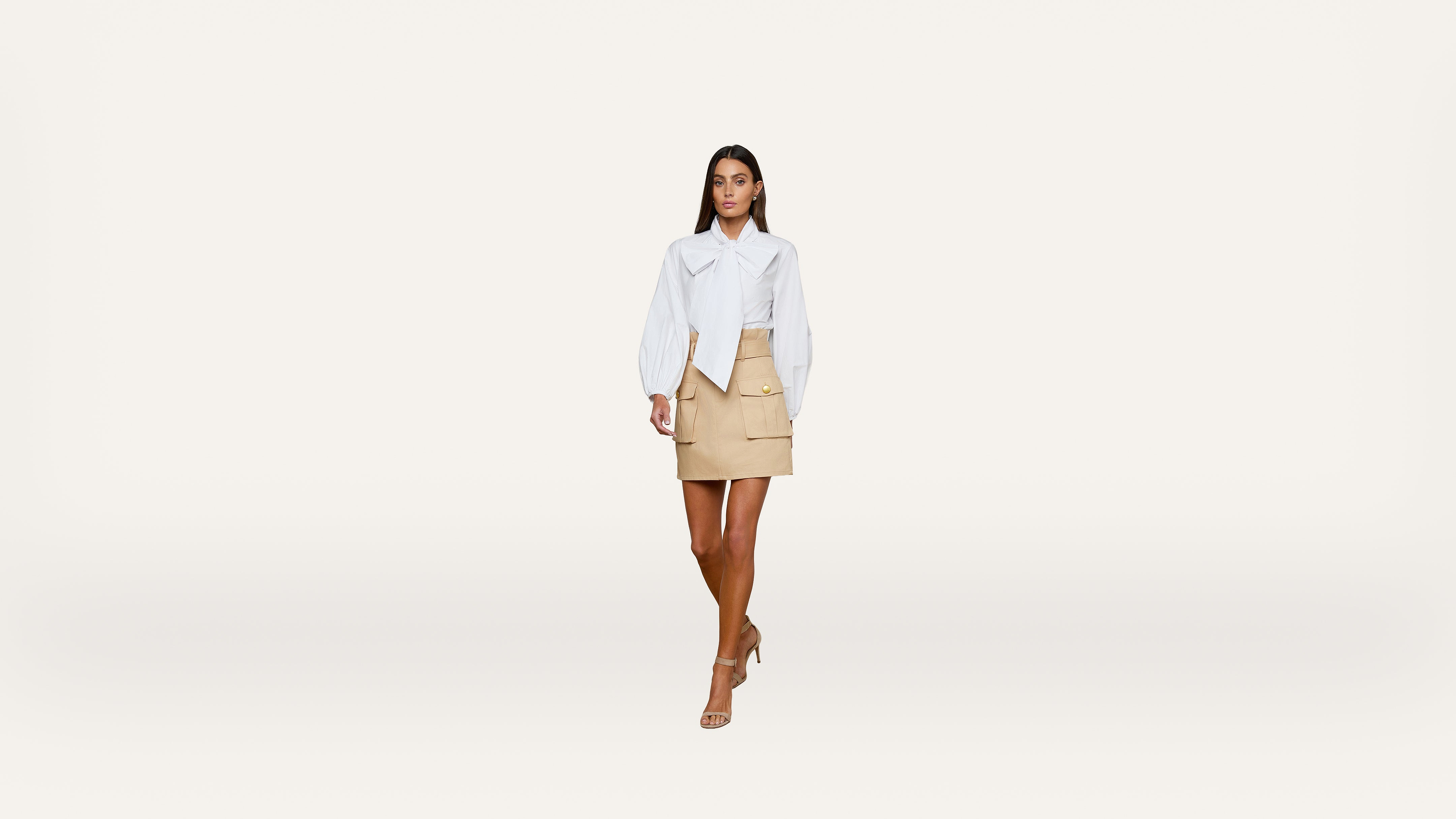 A woman walks forward wearing a white blouse, beige skirt, and nude heels against a plain white background.