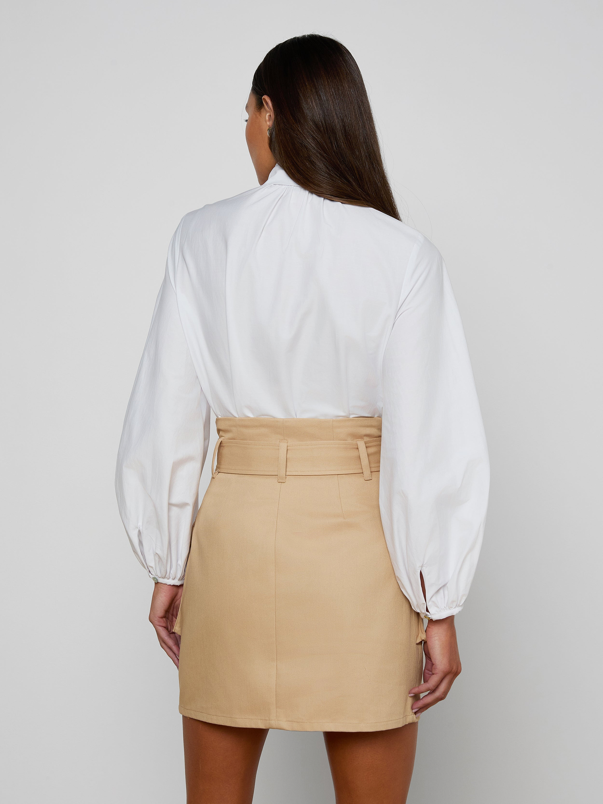 Seen from behind against a plain light background, a woman with long brown hair wears the Deon Tie-Neck Blouse—an organic cotton poplin white long-sleeve blouse with blouson sleeves—tucked into a high-waisted beige skirt.