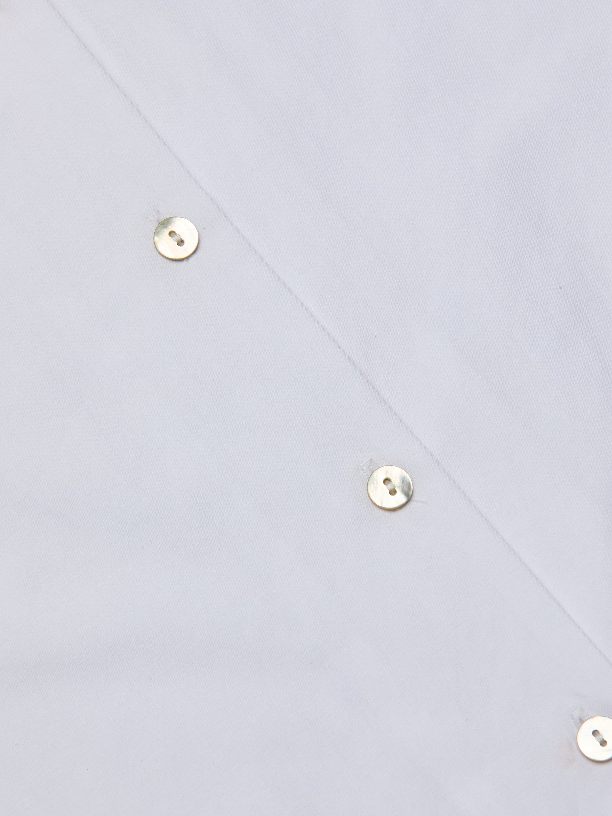 Close-up of the Deon Tie-Neck Blouse in white organic cotton poplin, featuring shiny metal snap buttons arranged diagonally, highlighting part of the garment. The reflective surface of the buttons adds a polished touch to the design.