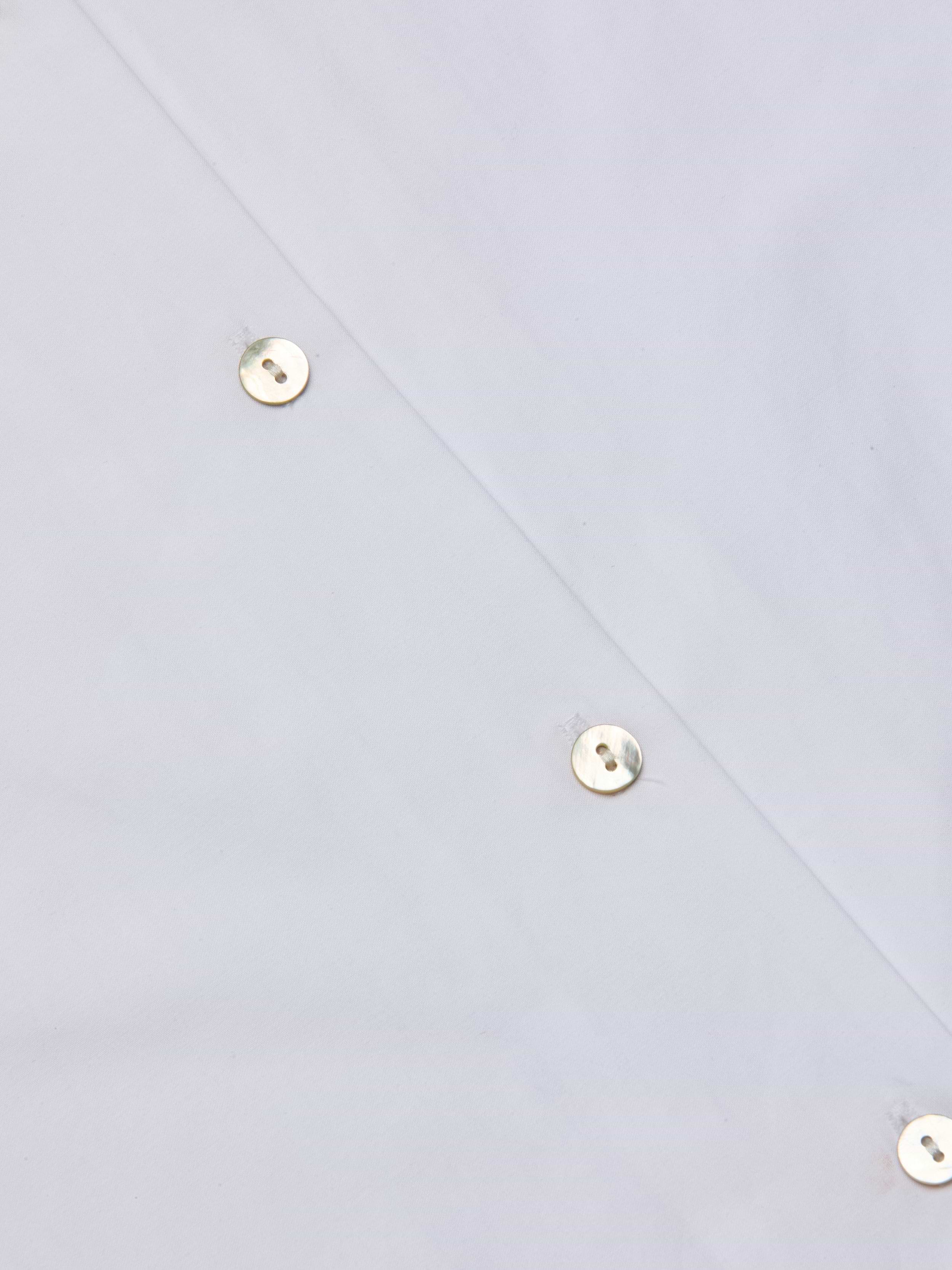 Close-up of the Deon Tie-Neck Blouse in white organic cotton poplin, featuring shiny metal snap buttons arranged diagonally, highlighting part of the garment. The reflective surface of the buttons adds a polished touch to the design.