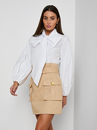 Wearing the Deon Tie-Neck Blouse in white with blouson sleeves, paired with a beige mini skirt featuring large front pockets and gold buttons, a woman stands against a plain light background.