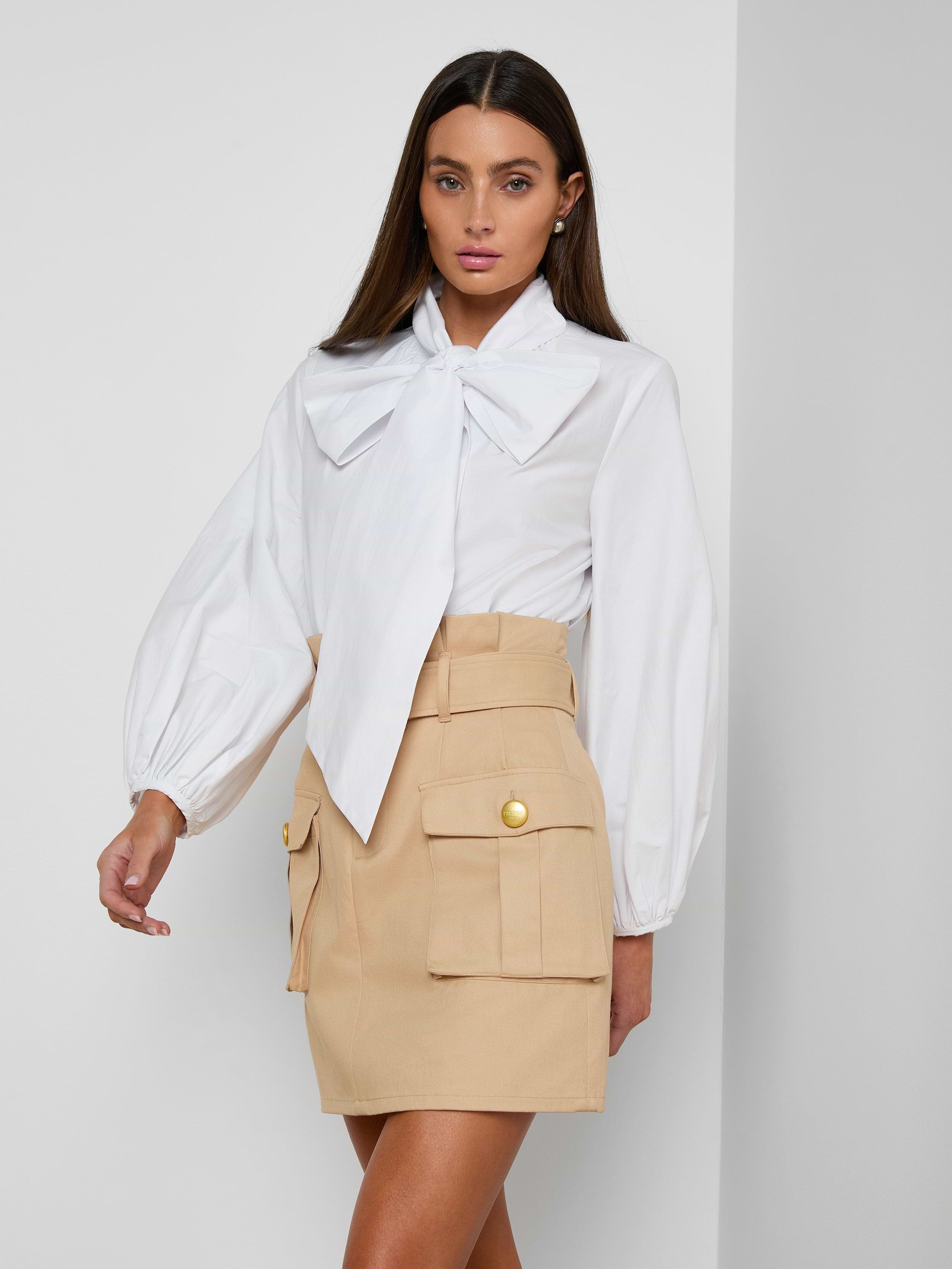 Wearing the Deon Tie-Neck Blouse in white with blouson sleeves, paired with a beige mini skirt featuring large front pockets and gold buttons, a woman stands against a plain light background.