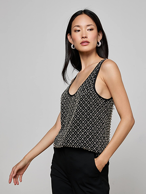 A woman with long black hair models the L’AGENCE Ziggy Crystal Tank and black pants, posing with one hand in her pocket against a plain gray background.
