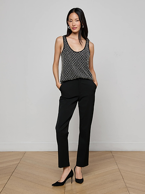 A woman stands indoors against a plain light wall, wearing the L'AGENCE Ziggy Crystal Tank with black pants and heels, her hands in her pockets and a slight smile.