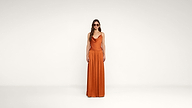 A woman stands against a plain white background, wearing sunglasses and an orange sleeveless dress with a loose skirt.