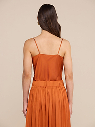 Una donna dai lunghi capelli castani è di spalle; indossa la canotta in raso Calista color arancio bruciato con spalline sottili, abbinata con eleganza a una gonna a pieghe dello stesso colore, su uno sfondo beige.
