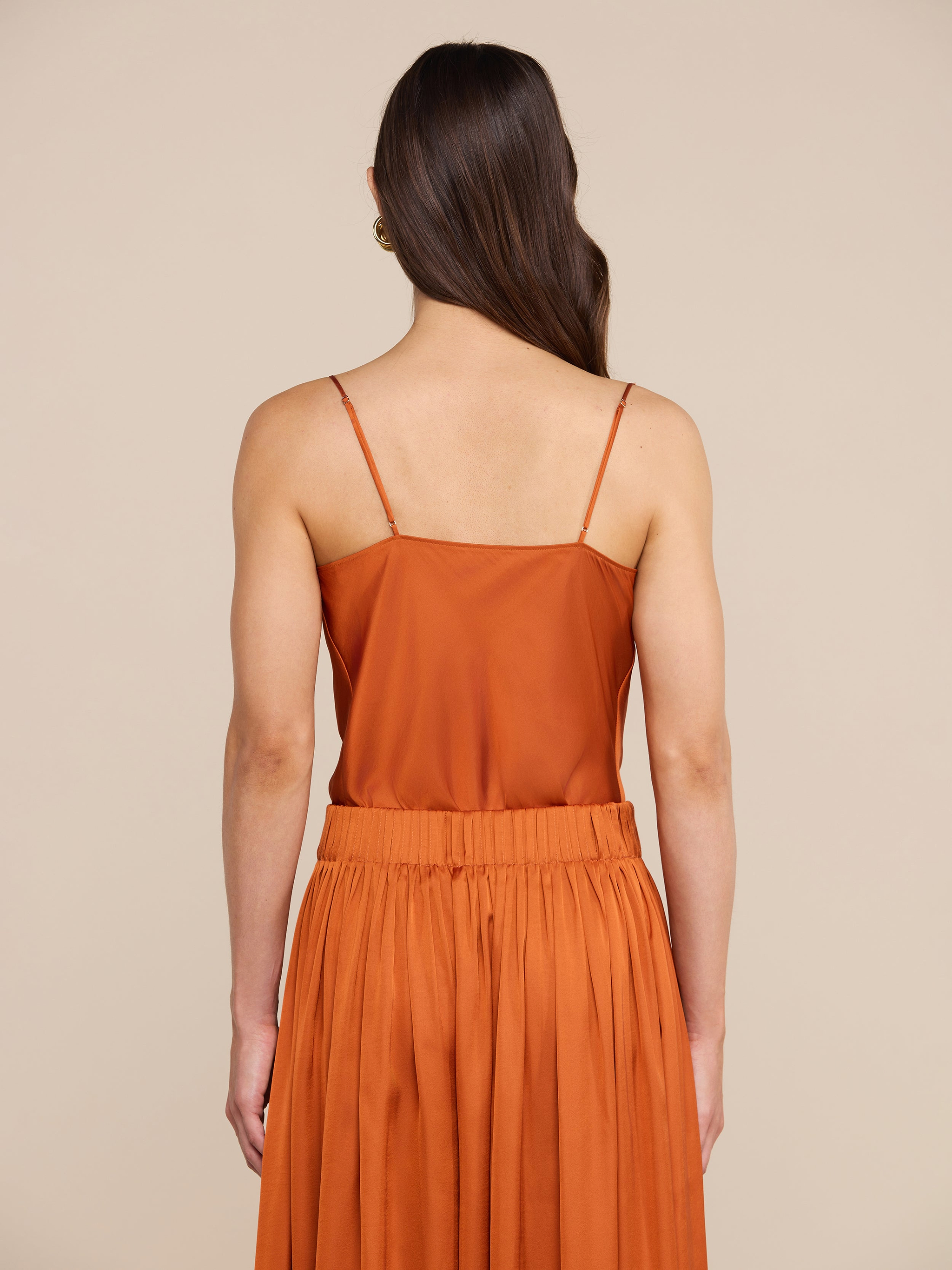 Una donna dai lunghi capelli castani è di spalle; indossa la canotta in raso Calista color arancio bruciato con spalline sottili, abbinata con eleganza a una gonna a pieghe dello stesso colore, su uno sfondo beige.