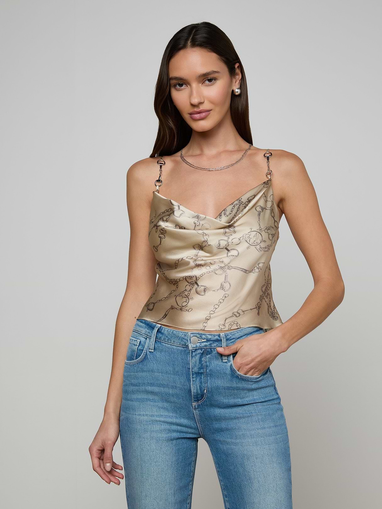A woman with long dark hair wears the Calista Silk Camisole, a cropped top featuring a horse bit chain print, paired with blue jeans and standing against a plain light background.