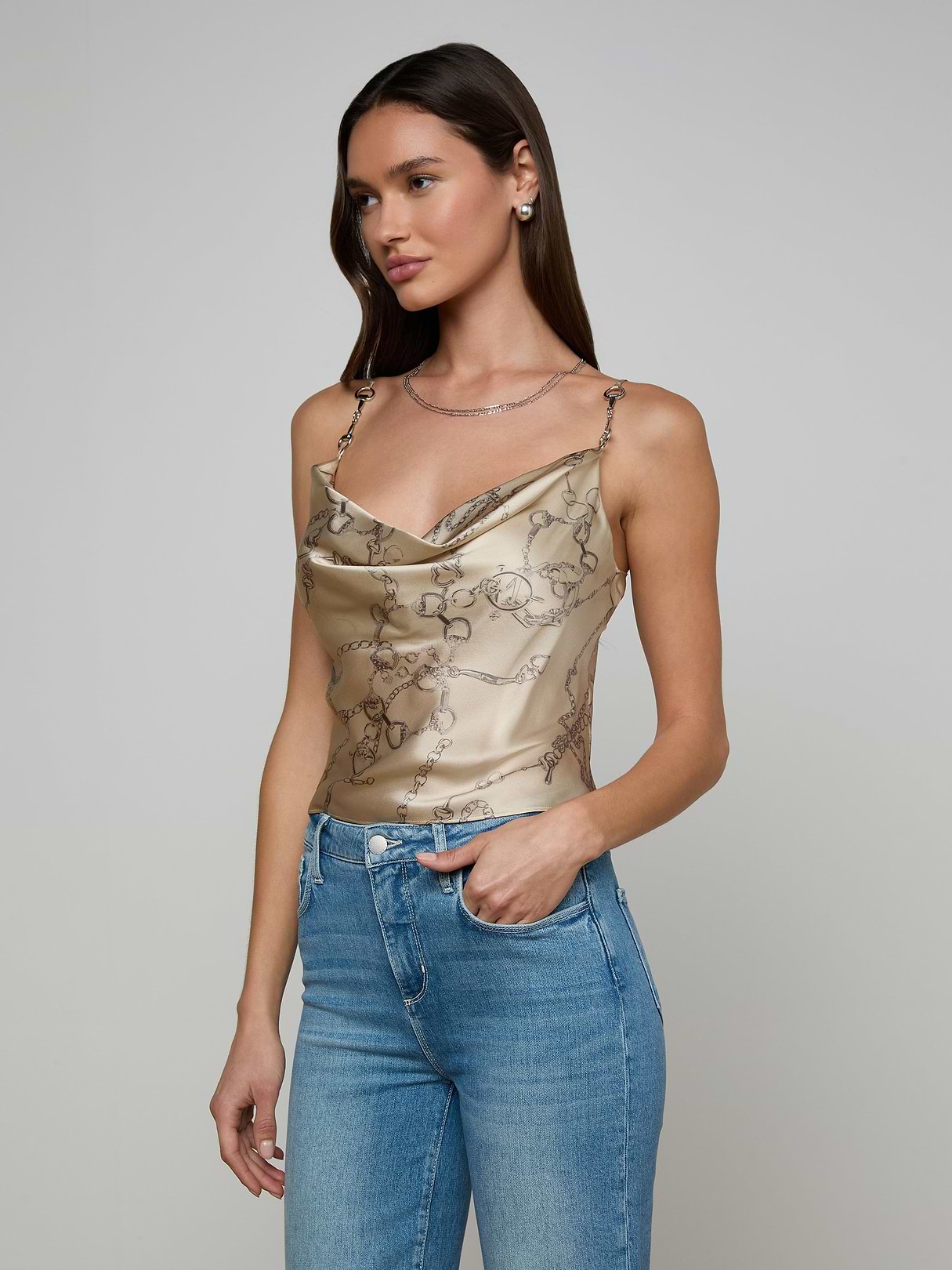 A woman wearing the Calista Silk Camisole, featuring a gold horse bit chain print, and blue jeans stands against an off-white background, looking to her right.