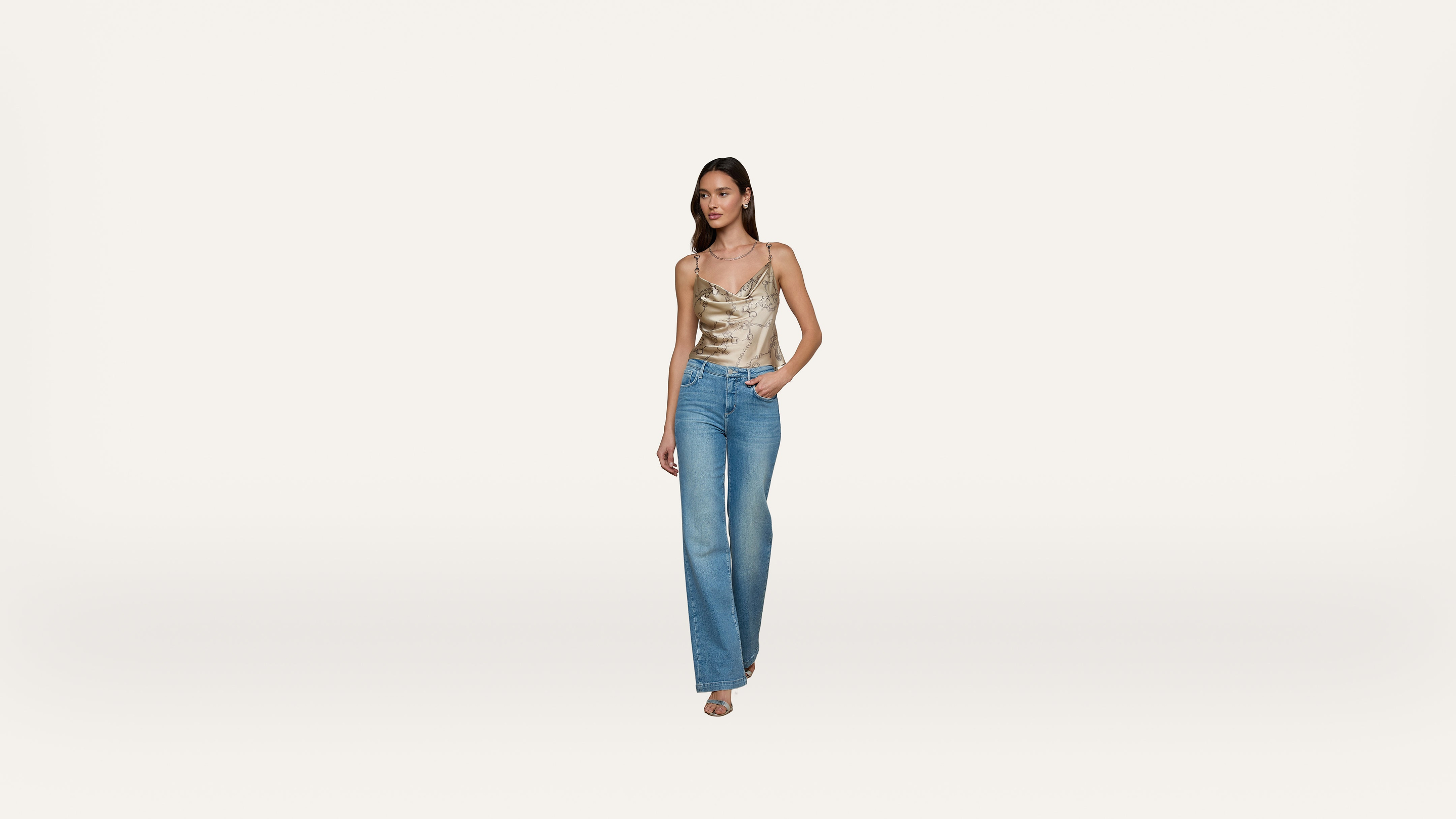 A woman wearing a gold satin camisole and blue jeans stands against a plain white background, with one hand in her pocket and the other by her side.