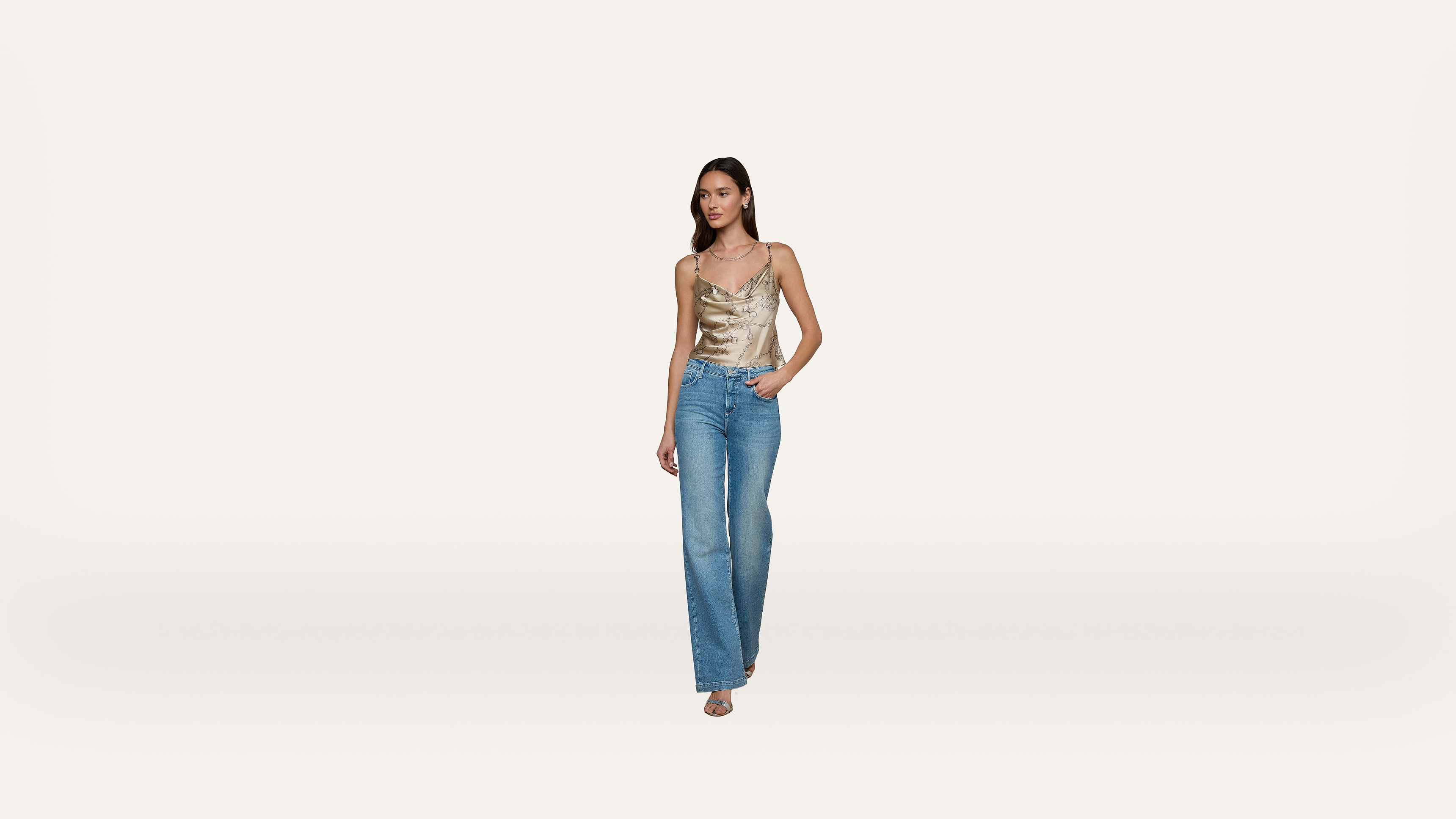 A woman wearing a gold satin camisole and blue jeans stands against a plain white background, with one hand in her pocket and the other by her side.