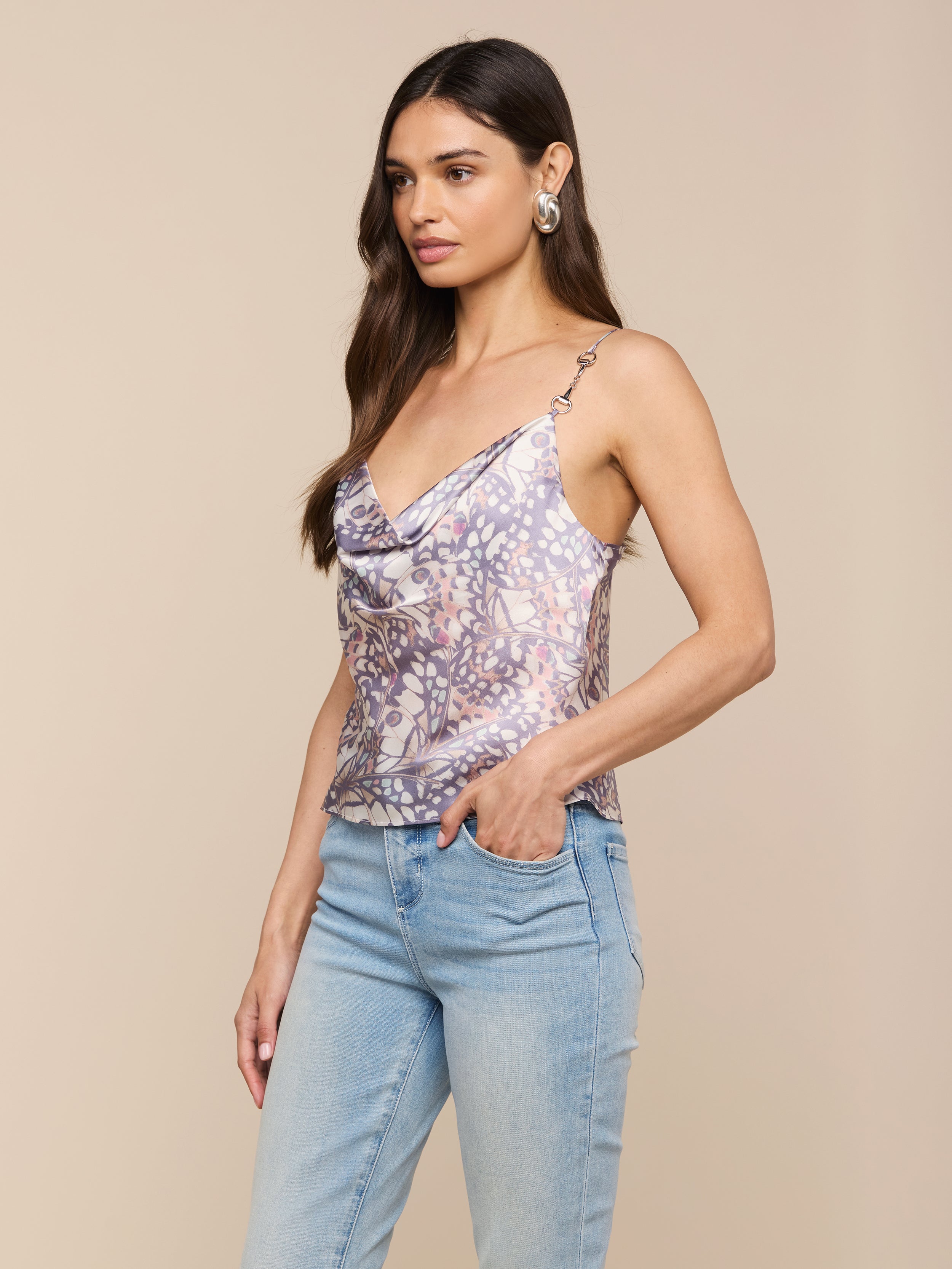 Woman with long brown hair wearing the Calista Satin Camisole—a floral-patterned, bias-cut satin sleeveless cowl-neck top—paired with light blue jeans, standing with one hand in her pocket against a plain background.