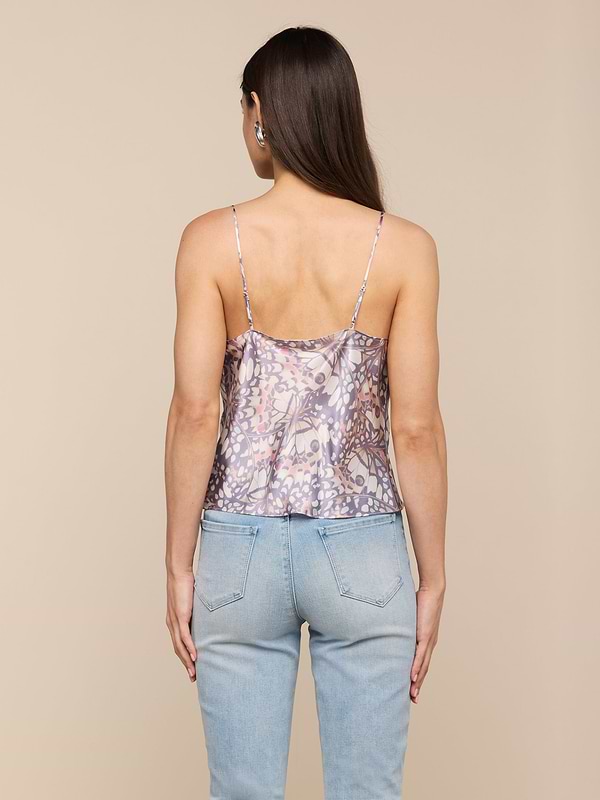 A woman with long brown hair stands facing away, wearing the Calista Satin Camisole featuring a butterfly wing print paired with light blue jeans against a plain, light background.