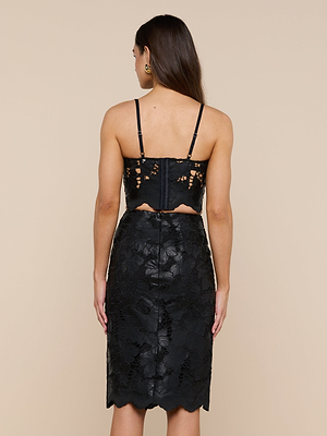 Une femme aux longs cheveux bruns porte le Sonya Vegan Leather Bustier avec des bretelles réglables et une jupe taille haute assortie, vue de dos sur un fond beige uni.