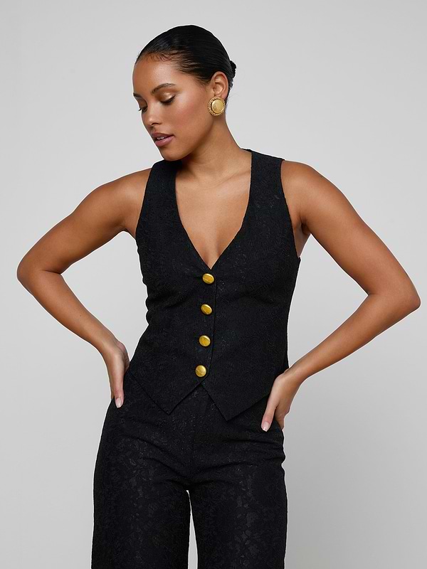 A woman models the Kino Lace Vest—a sleeveless black vest with gold buttons and modern tailoring—paired with matching black pants, posing with hands on hips against a plain, light background.