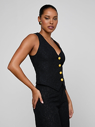 A woman poses confidently against a plain light background, wearing the Kino Lace Vest—a sleeveless black lace vest with gold buttons—paired with matching pants. She stands with one hand on her hip, highlighting the modern tailoring of the look.