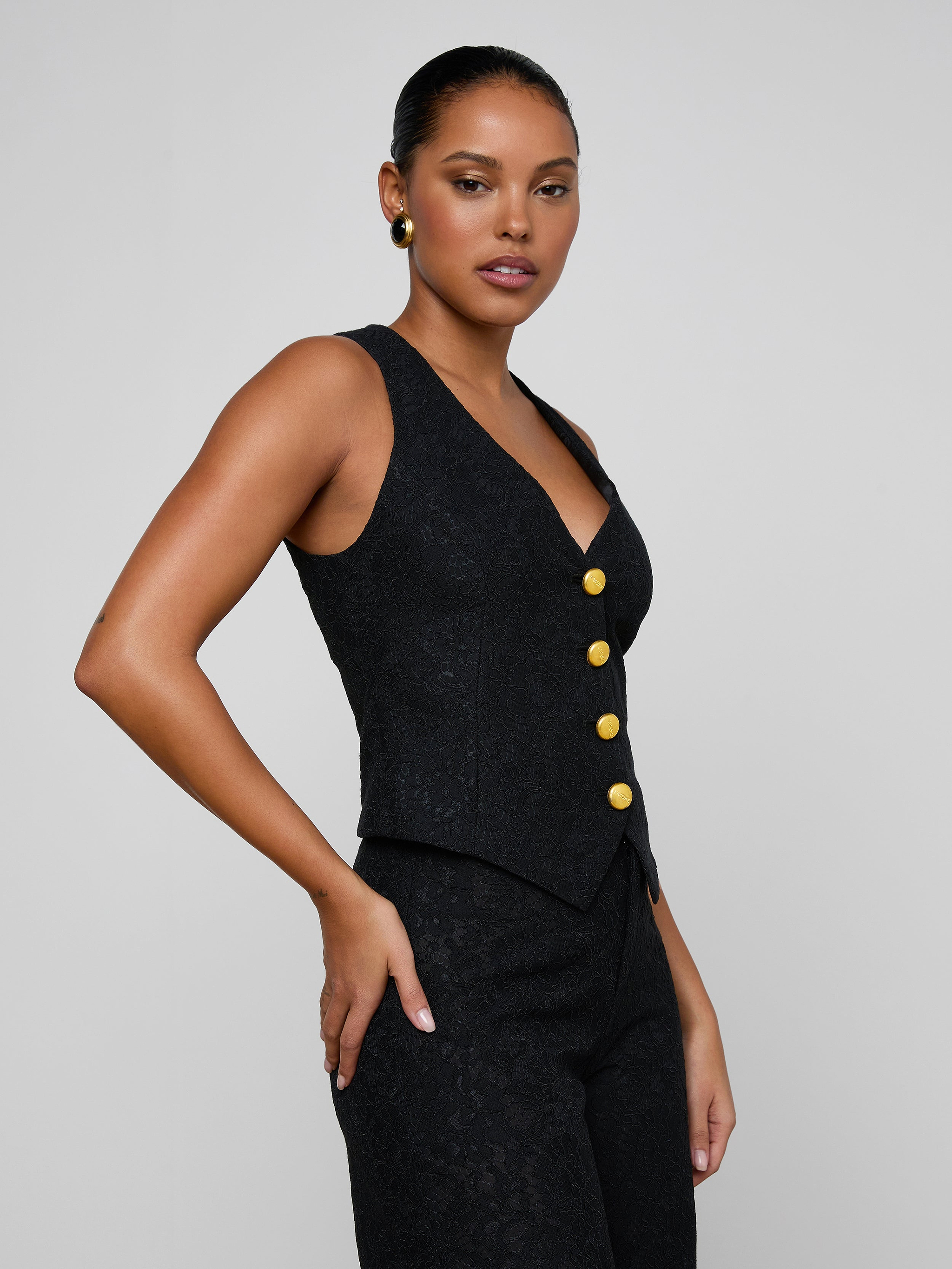 A woman poses confidently against a plain light background, wearing the Kino Lace Vest—a sleeveless black lace vest with gold buttons—paired with matching pants. She stands with one hand on her hip, highlighting the modern tailoring of the look.