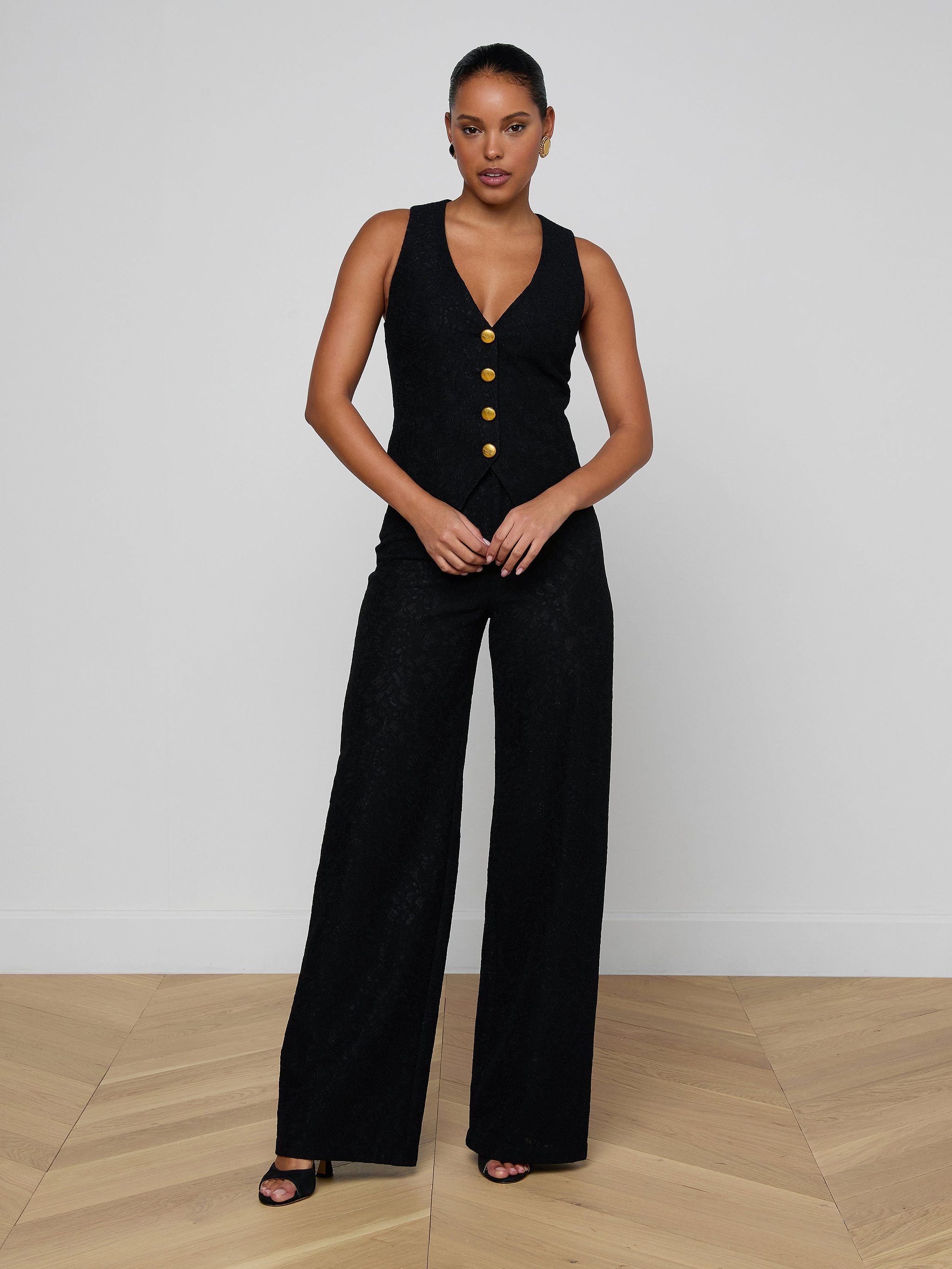 A woman stands against a plain light background, wearing the Kino Lace Vest—a sleeveless black piece featuring modern tailoring and gold buttons—paired with black heeled sandals. She faces forward, her hands clasped in front of her.