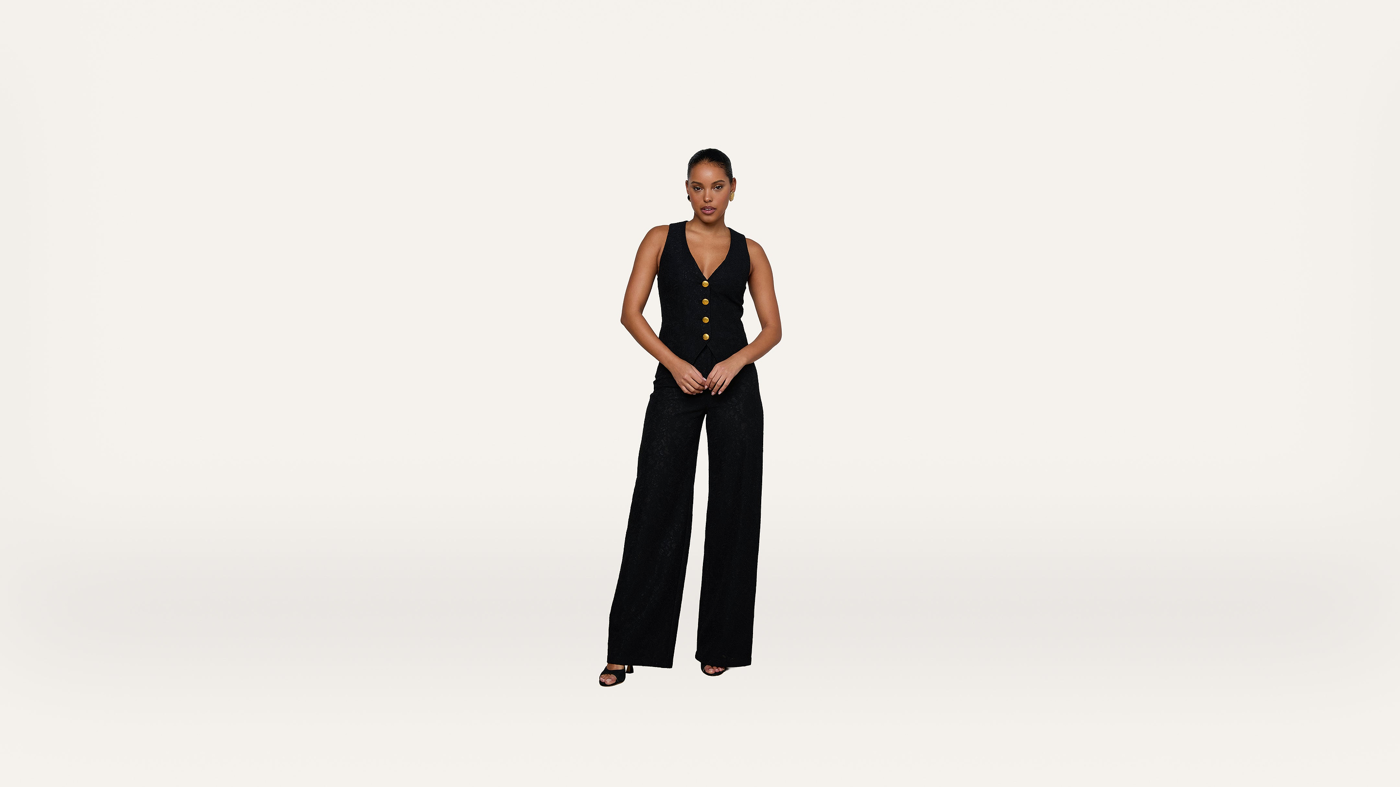 A woman stands against a plain white background, wearing a sleeveless black vest with buttons and wide-leg black pants, paired with black heels.