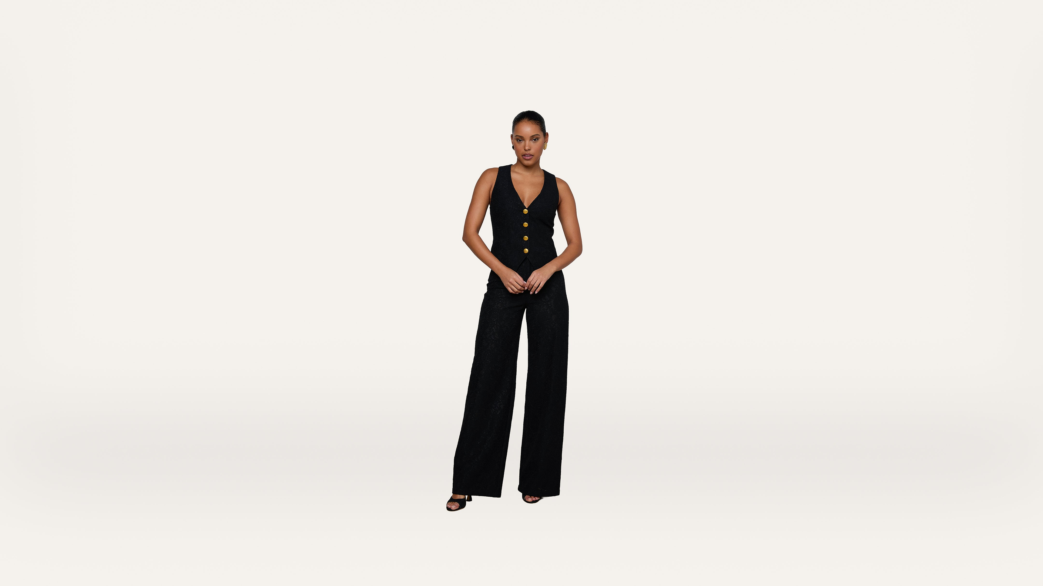 A woman stands against a plain white background, wearing a sleeveless black vest with buttons and wide-leg black pants, paired with black heels.
