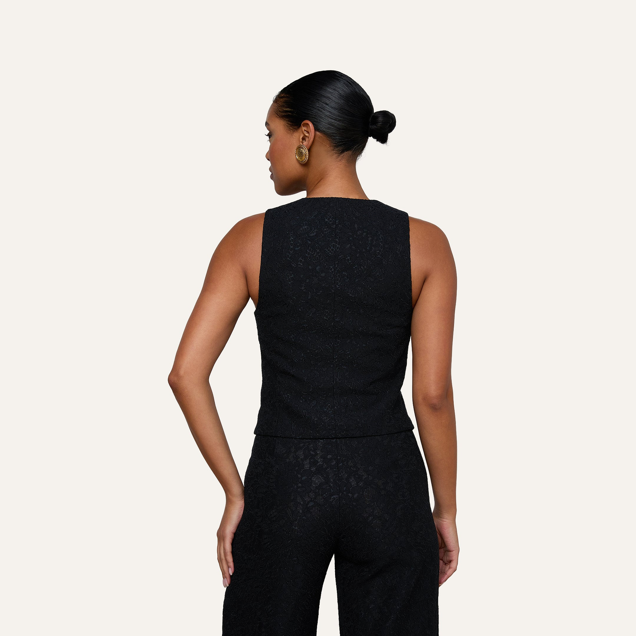 Woman with hair in a bun wearing the Kino Lace Vest—a sleeveless black top with gold buttons—and matching black pants, shown from the back against a plain off-white background.