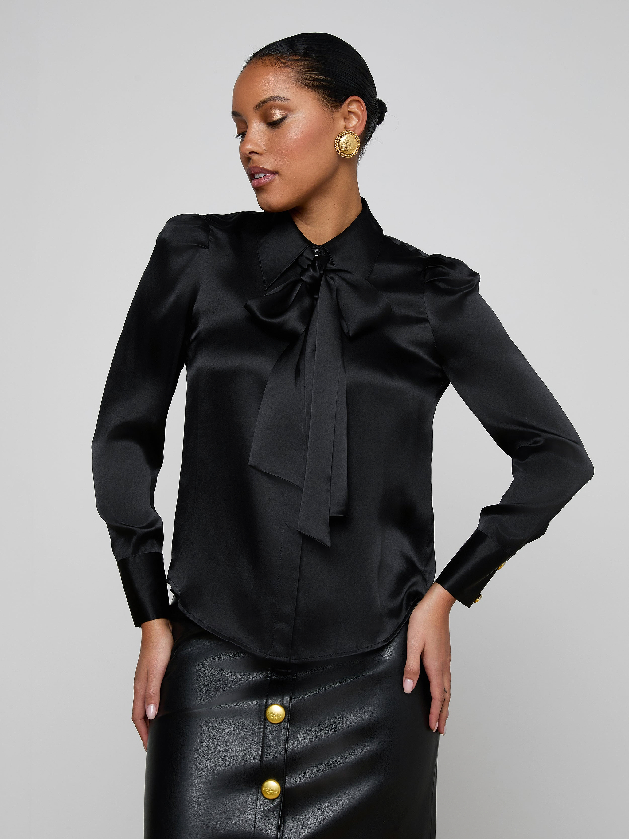Wearing the Honey Silk Blouse, a woman poses with one hand on her hip. The black silk blouse features a bow tie detail and is complemented by gold button earrings. She pairs it with a coordinating black skirt adorned with large gold buttons, standing against a plain light background.