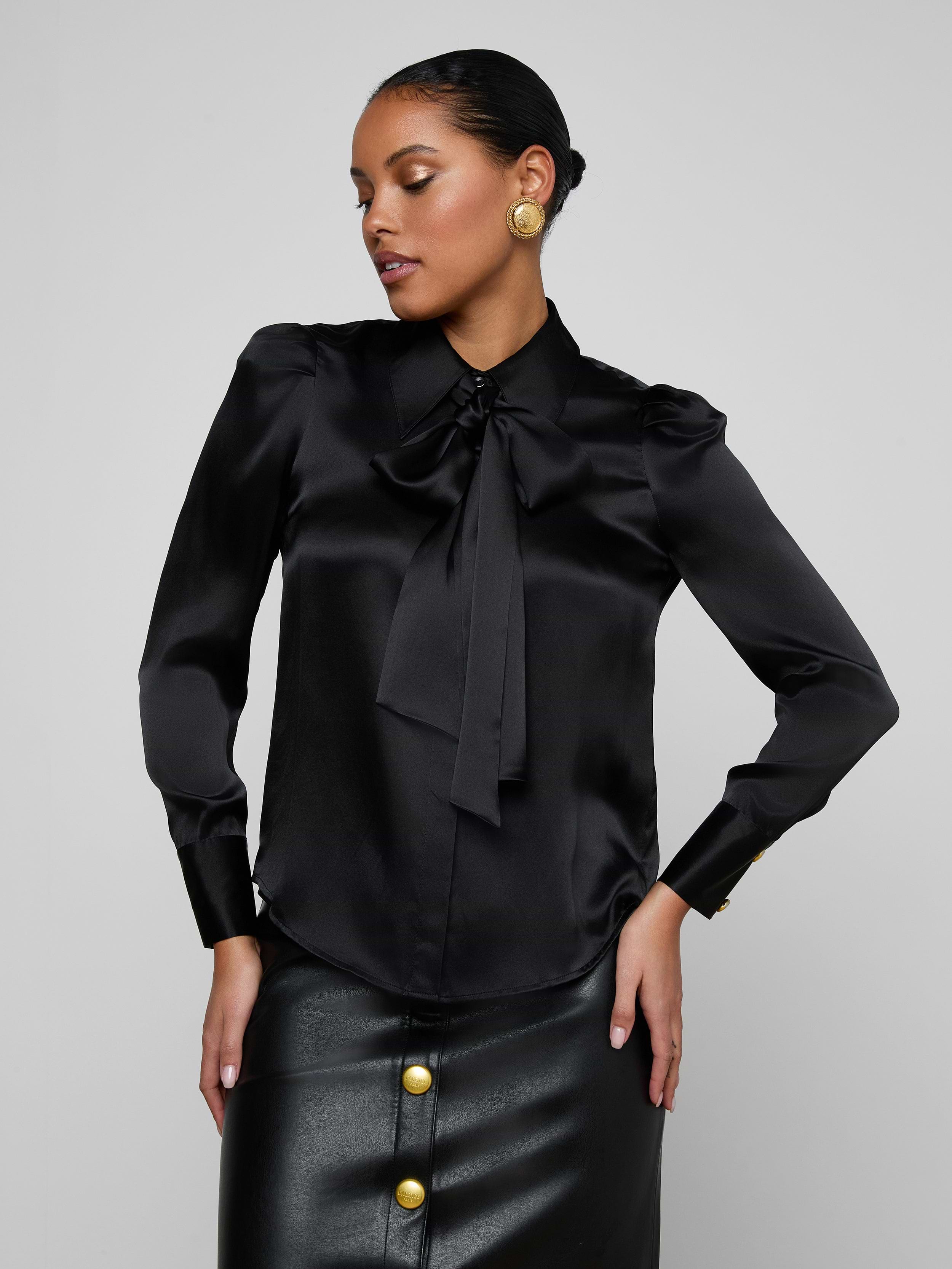 Wearing the Honey Silk Blouse, a woman poses with one hand on her hip. The black silk blouse features a bow tie detail and is complemented by gold button earrings. She pairs it with a coordinating black skirt adorned with large gold buttons, standing against a plain light background.