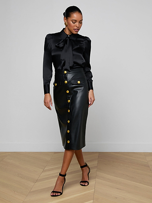 A woman wearing the Honey Silk Blouse with a bow collar and a high-waisted black leather midi skirt with gold buttons, paired with black strappy heels, stands on a wooden floor against a plain white wall.
