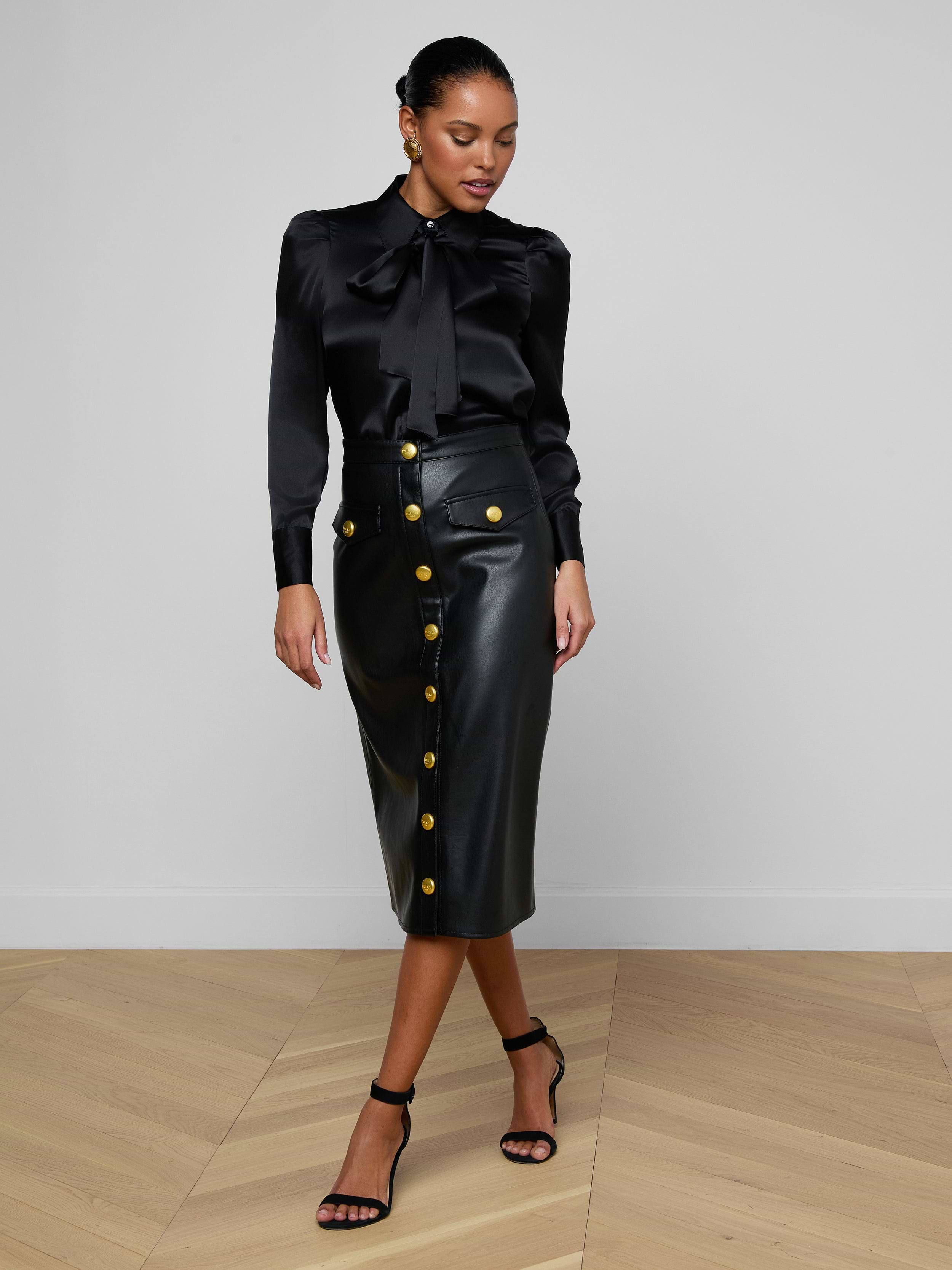 A woman wearing the Honey Silk Blouse, paired with a black leather skirt featuring gold buttons and black heels, stands against a plain white background.