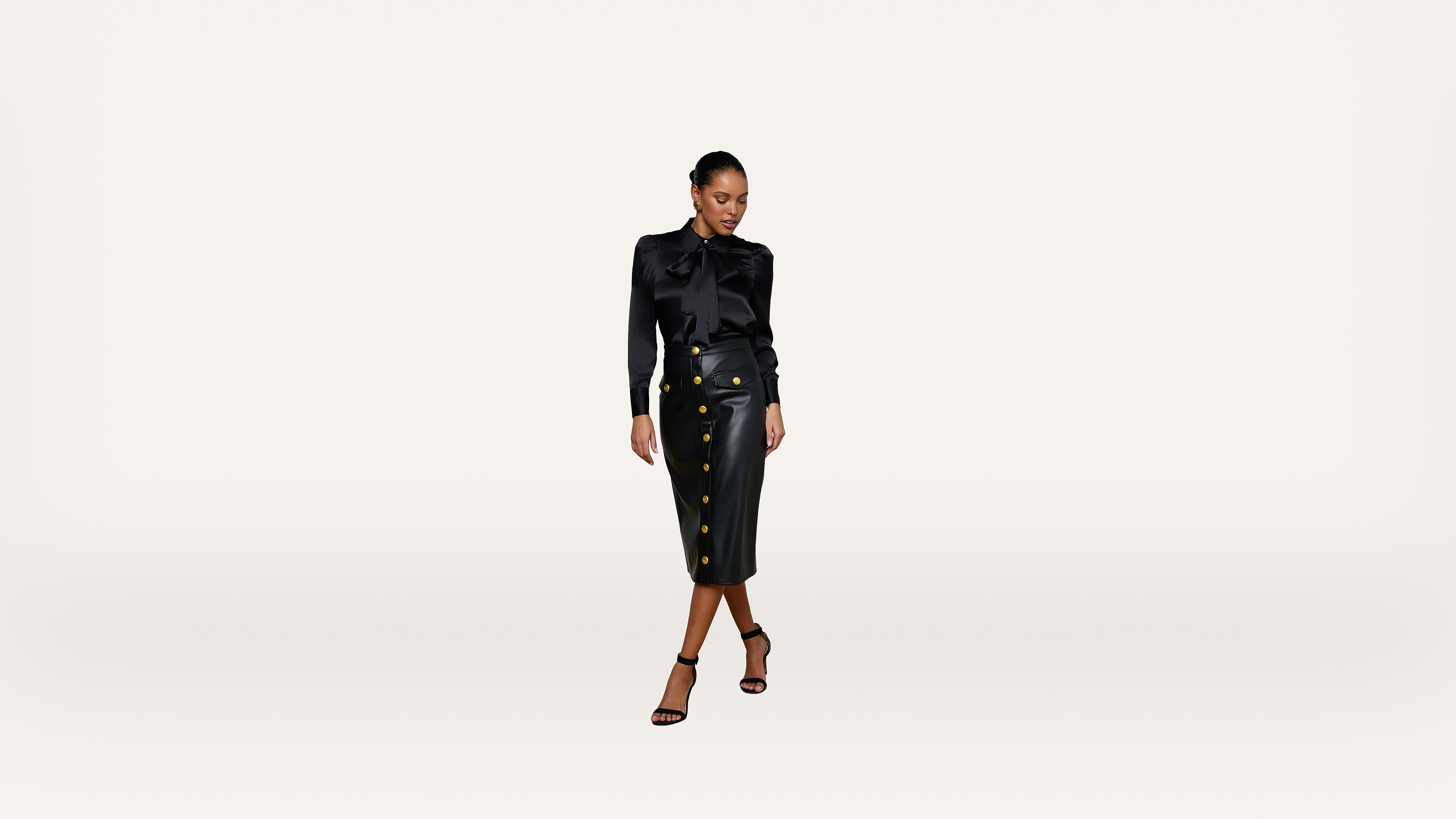 A woman in a black blouse and a high-waisted black skirt with gold buttons stands against a plain white background, looking down and slightly to the side.