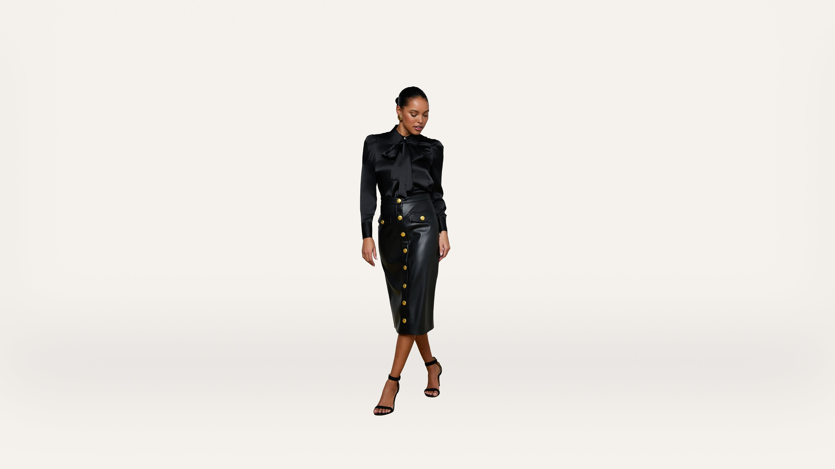 A woman in a black blouse and a high-waisted black skirt with gold buttons stands against a plain white background, looking down and slightly to the side.