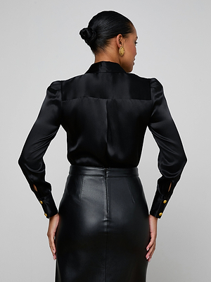 A woman with her hair in a low bun faces away, wearing the Honey Silk Blouse—a sleek black silk top with long sleeves and gold buttons—paired with a high-waisted black leather skirt.