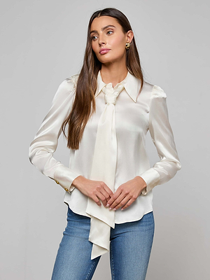 A woman with long brown hair wears the Honey Silk Blouse, featuring a tie neck and gold button details, paired with blue jeans against a plain light gray background.