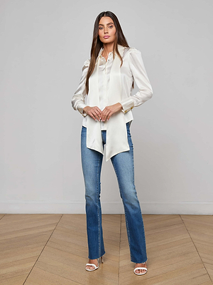 A woman with long brown hair stands indoors on a wooden floor, facing the camera with a neutral expression. She wears the Honey Silk Blouse, blue jeans, and white heeled sandals against a plain backdrop.