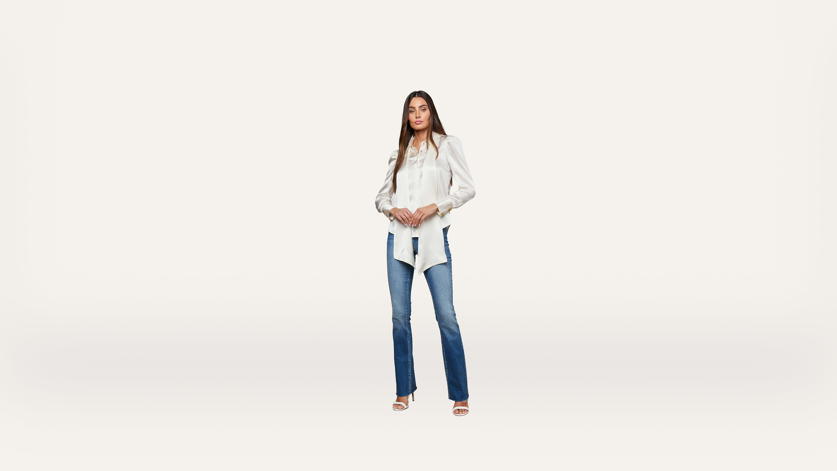 A woman stands facing forward, wearing a white blouse with puffed sleeves, blue jeans, and open-toed shoes against a plain white background.