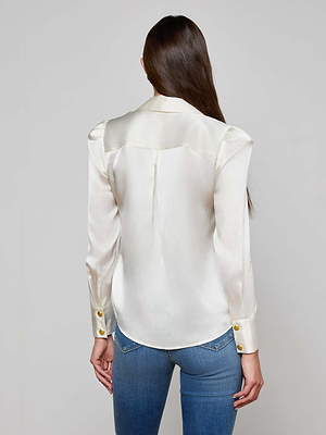 A woman with long brown hair, seen from behind, wears the Honey Silk Blouse—a shiny cream-colored silk charmeuse top with gold cuff buttons—paired with blue jeans against a plain light gray background.