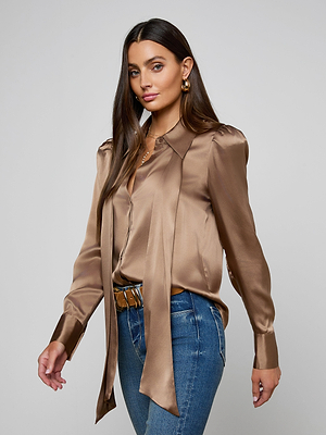 A woman with long brown hair wears the Honey Silk Blouse—a shiny bronze silk charmeuse tie-neck top—with blue jeans. She stands against a plain gray background, looking confidently at the camera.