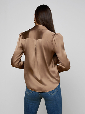 A woman with long brown hair is shown from the back, wearing the Honey Silk Blouse— a shiny brown charmeuse top with puffed shoulders— and blue jeans, standing against a plain light gray background.