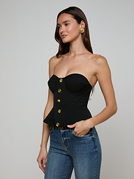 A woman wearing the Anna Corset Top—a high-fashion, strapless black corset accented with gold buttons—and blue jeans stands with one hand in her pocket against a plain light background.
