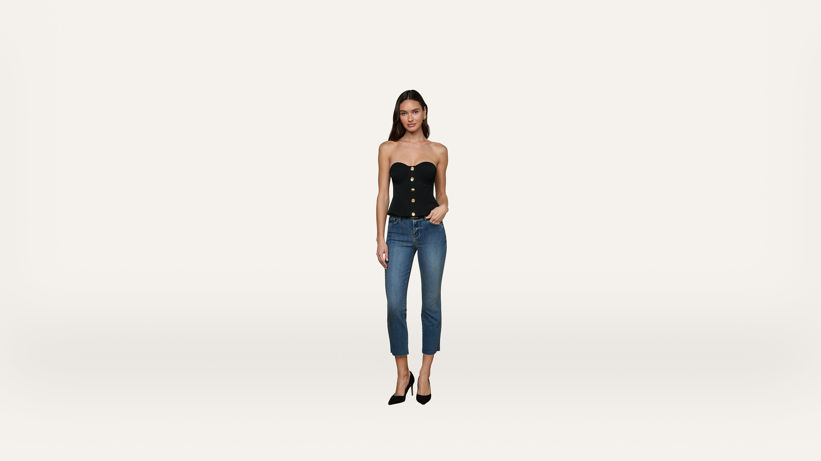 A woman wearing a black strapless top with buttons, blue cropped jeans, and black heels stands against a plain white background.