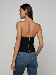 Wearing the Anna Corset Top—a high-fashion going-out piece with a sculptural silhouette—the woman faces away, highlighting the strapless black design with a visible gold zipper and paired with blue jeans. Her straight, long hair is styled to showcase large gold hoop earrings, set against a plain off-white background.