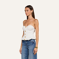 Una donna dai lunghi capelli castani e con orecchini a cerchio dorati posa su uno sfondo beige indossando l’Anna Corset Top, un top bianco senza spalline in stile peplum con bottoni dorati, abbinato a jeans blu e a un braccialetto dorato per un look chic e di alta moda.