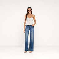 Indossando il top a corsetto Anna, una donna posa con jeans blu a gamba larga, sandali bianchi con tacco, occhiali da sole e un braccialetto d’oro su uno sfondo beige a tinta unita.
