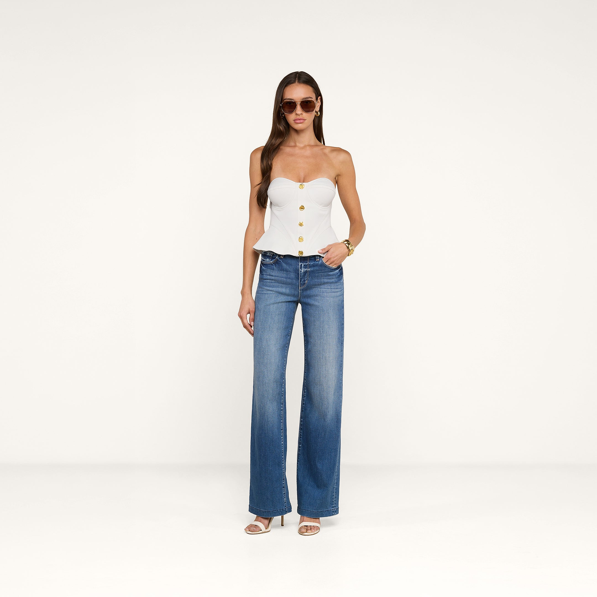 Indossando il top a corsetto Anna, una donna posa con jeans blu a gamba larga, sandali bianchi con tacco, occhiali da sole e un braccialetto d’oro su uno sfondo beige a tinta unita.