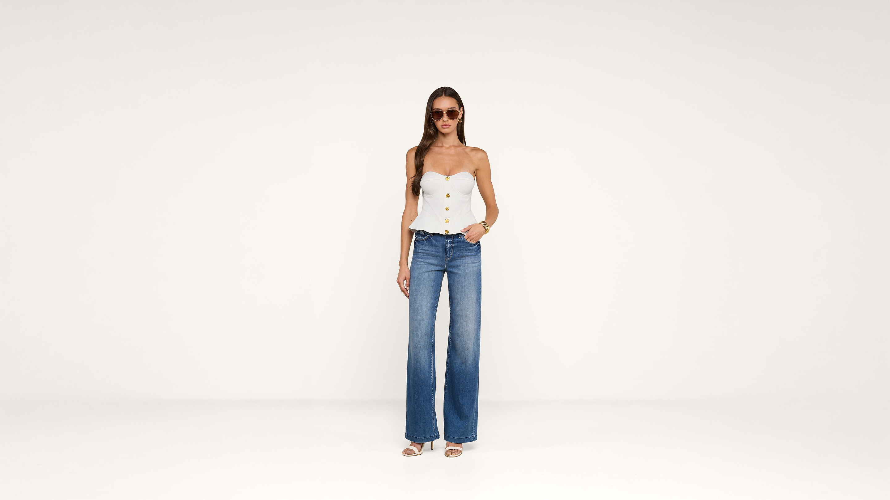 Woman wearing sunglasses, a white strapless top with buttons, blue wide-leg jeans, and white sandals, standing against a plain white background.